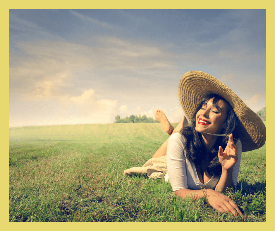 Uśmiechnięta kobieta, która ma kapelusz na głowie i leży na łące. Widać, że jest na wakacjach.